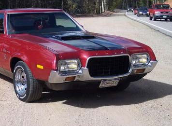 1971 Ford Ranchero - A red pickup truck from the 1970s is seen driving on a road, with other vehicles in the background.