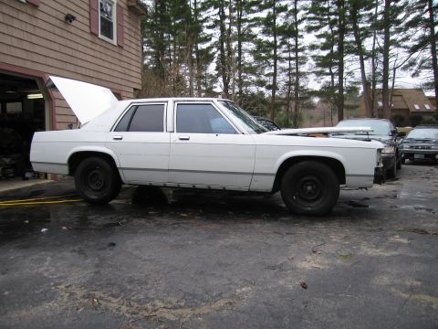 1991 Ford LTD Crown Victoria - A white sedan sits in a driveway with its hood and trunk open, parked near other vehicles.