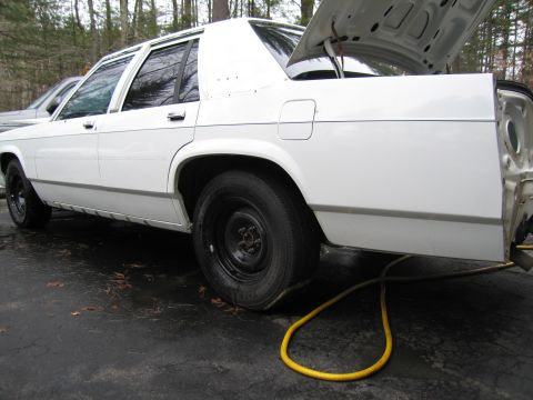 1991 Ford LTD Crown Victoria - A white sedan sits with its trunk open and a flat tire, revealing some damage to the rear.