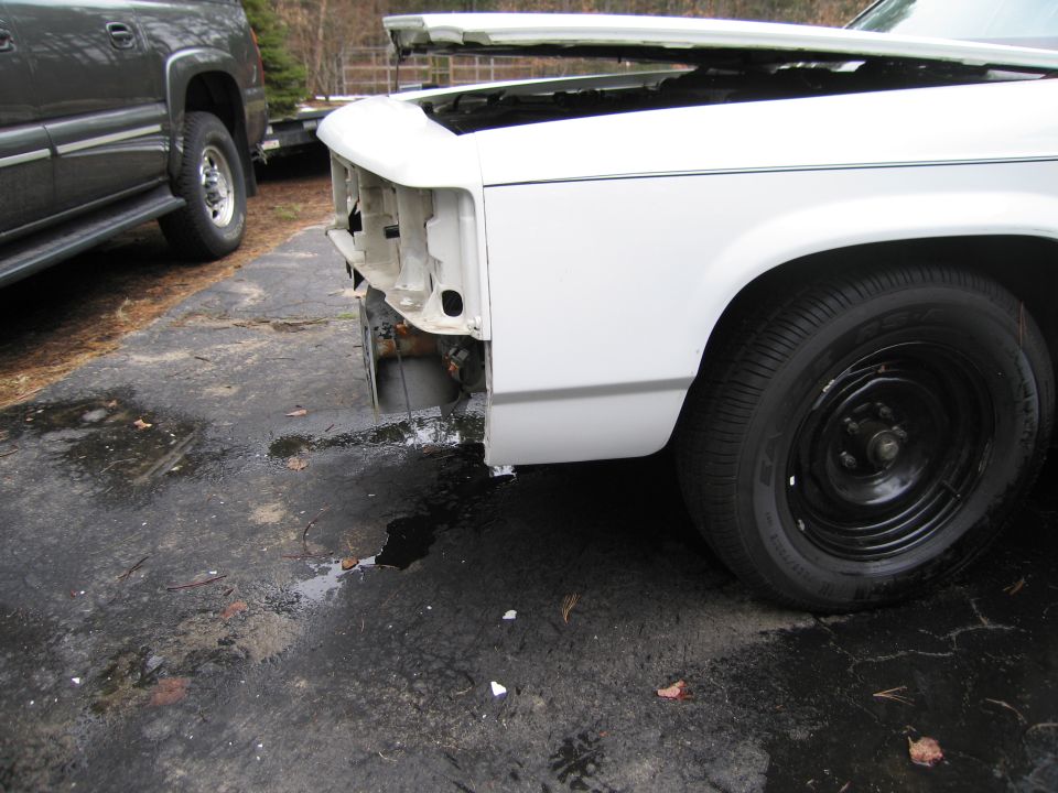 White car front end, hood open, missing grille and headlights. Another vehicle is parked nearby