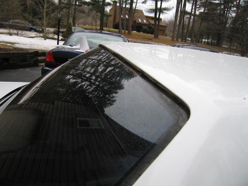 White car roof and rear window with reflection of trees and a house; another dark car is visible behind it
