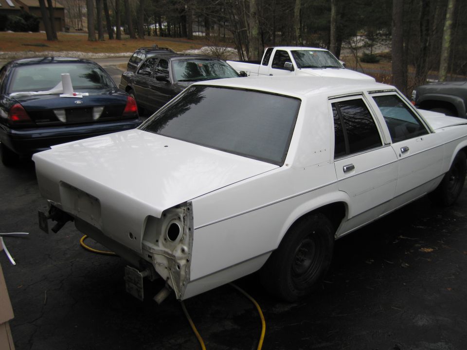 White sedan with rear bumper removed, parked outside. Other cars and a truck are visible in the background