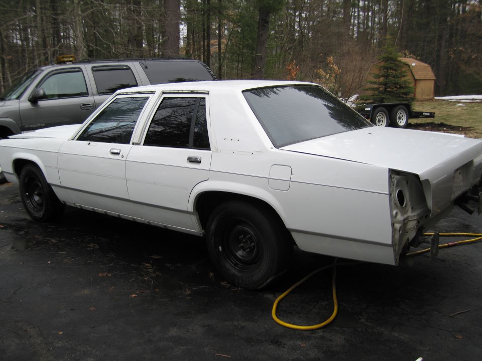 White sedan with black wheels, missing rear bumper and taillights, parked on asphalt