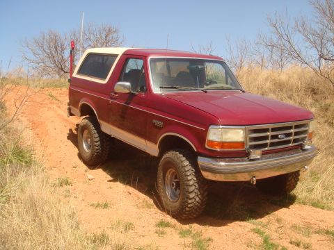 1992 Ford Bronco - A red 1992 Ford Bronco with a white removable top is shown off-road on a dirt path.