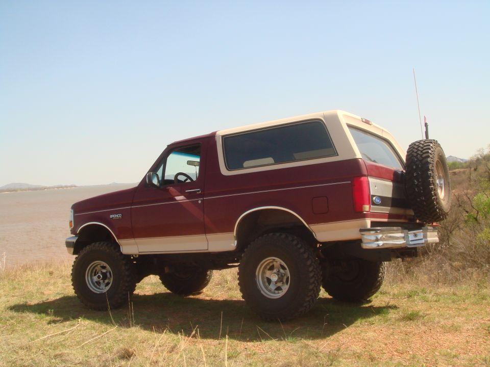 Maroon and tan Ford Bronco parked on grassy hill near water. Spare tire mounted on rear