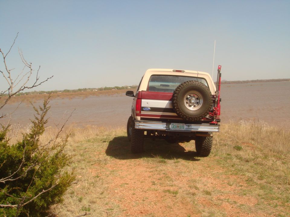 1992 Ford Bronco, rear view, red and tan, with spare tire mounted on back, parked on grassy bank near water