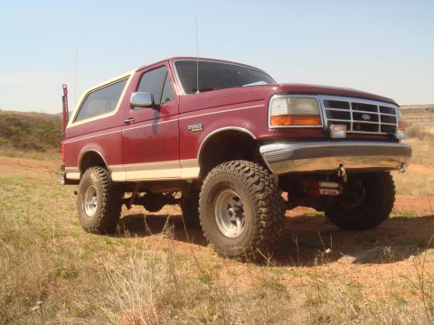 1992 Ford Bronco - Lifted burgundy Ford Bronco with a tan/cream removable hardtop and lower body stripe, posed at a three-quarter front angle on red dirt and…
