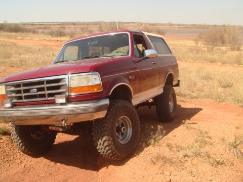 1992 Ford Bronco - Maroon Ford Bronco with white top and lifted suspension drives on a dirt road.