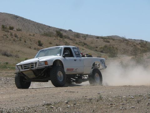 1990 Ford Bronco - white off-road race truck lifts its front wheels off a dirt hillside trail, trailing dust in the desert.
