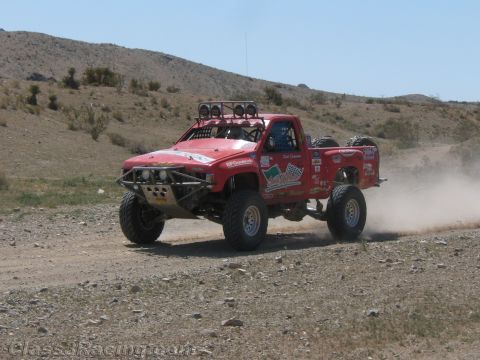 1990 Ford Bronco - red off-road race truck fitted with a roof light bar speeds along a rocky desert hillside trail.