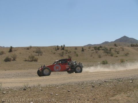 1990 Ford Bronco - red single-seat tube-frame off-road race buggy speeds along a dirt desert road with dust behind it.