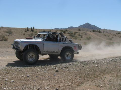 1990 Ford Bronco - silver early Ford Bronco off-road racer numbered 348 drives a dirt desert road below a pointed mountain peak.