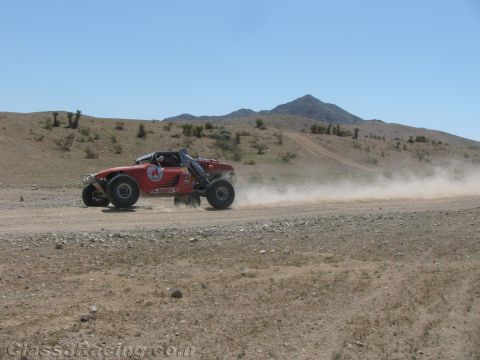 1990 Ford Bronco - red tube-frame off-road race buggy drives along a dirt desert road with a dust trail and a peak behind it.