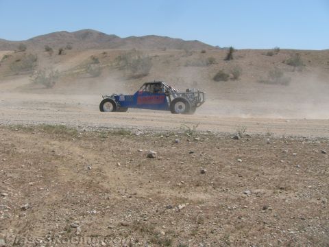 1990 Ford Bronco - blue and red tube-frame off-road race buggy races across a wide desert wash, raising a long cloud of dust.