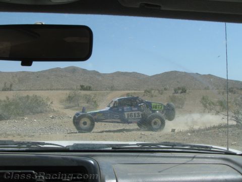 1990 Ford Bronco - Number 1613 single-seat off-road race buggy speeds across a dusty desert course, viewed through a vehicle windshield.