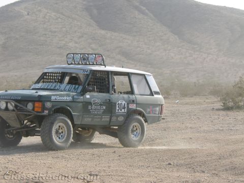1990 Ford Bronco - dark green Range Rover off-road race truck numbered 304, fitted with a roof light bar and racing decals, sits in the desert.