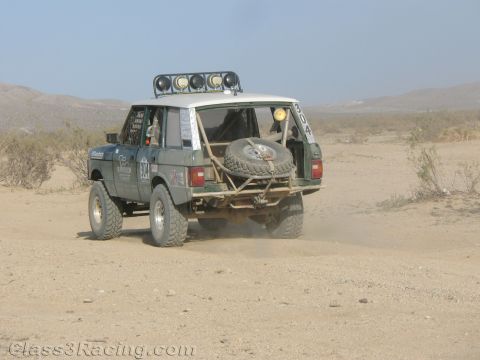1990 Ford Bronco - Rear three-quarter view of a green off-road Range Rover race truck with roof light bar and rear-mounted spare tire driving on a dusty deser…