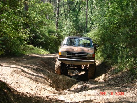 1987 Ford Bronco - 1987 Ford Bronco navigates a challenging muddy off-road course surrounded by trees.
