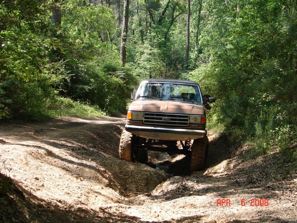 1987 Ford Bronco driving through mud on a wooded trail. "Monster Performance" sticker on windshield