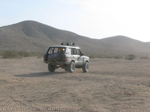 1990 Ford Bronco - silver off-road Range Rover with roof light bar and rear spare tire sits on an open desert flat with hills behind.