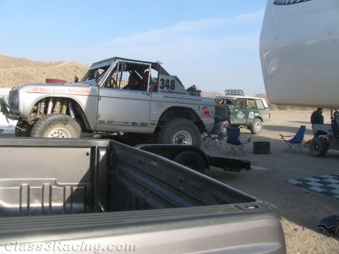 1990 Ford Bronco - silver tube-framed early Ford Bronco race truck numbered 348 sits in a desert pit area, viewed over the bed of a pickup.