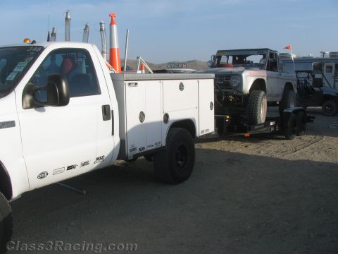 1990 Ford Bronco - white utility-bed service pickup parked next to a trailer carrying a silver early Ford Bronco race truck at a desert event.