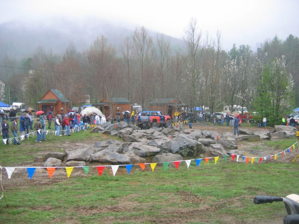 Spectators watch a red lifted 4x4 climb boulders at a muddy off-road event