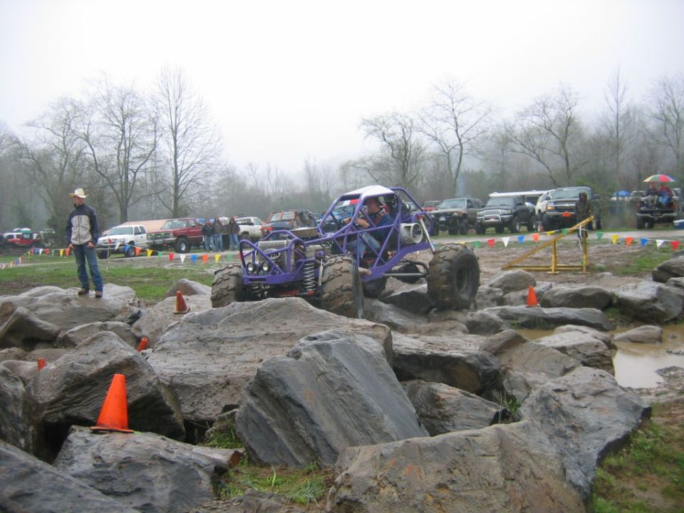 Purple tube-frame rock buggy climbing wet boulders at a foggy off-road crawling course