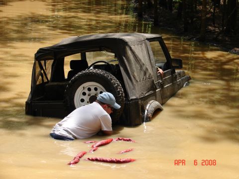 1987 Ford Bronco - Off-road vehicle stuck in muddy water with a person trying to get it out.