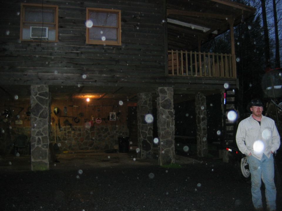 Man in jeans and a light jacket stands in front of a stilted wooden house at dusk
