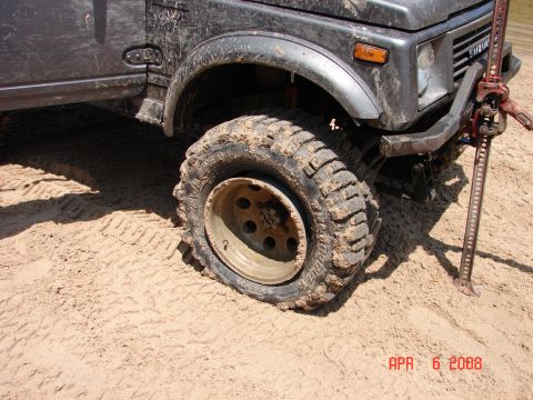 1987 Ford Bronco - A mud-covered Suzuki Samurai is shown with a flat tire and a Hi-Lift jack attached to the front bumper.