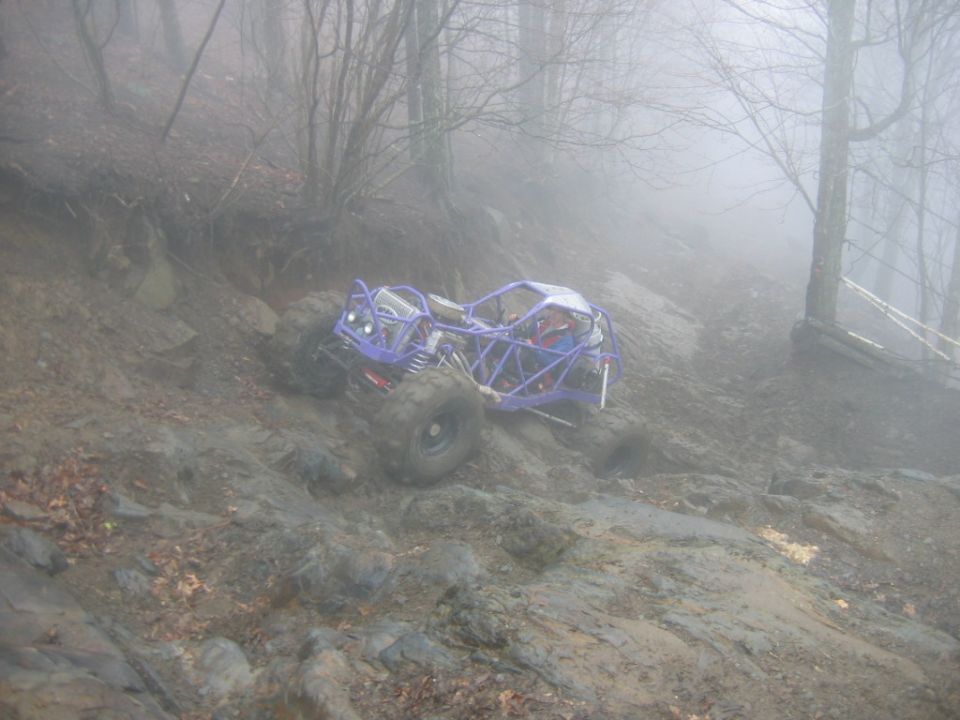 Purple off-road buggy with roll cage climbing a steep, muddy, rocky incline in a foggy forest