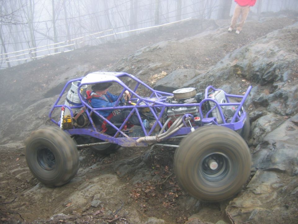 Purple off-road buggy with large tires climbing a rocky incline, driver wearing a blue jacket and helmet