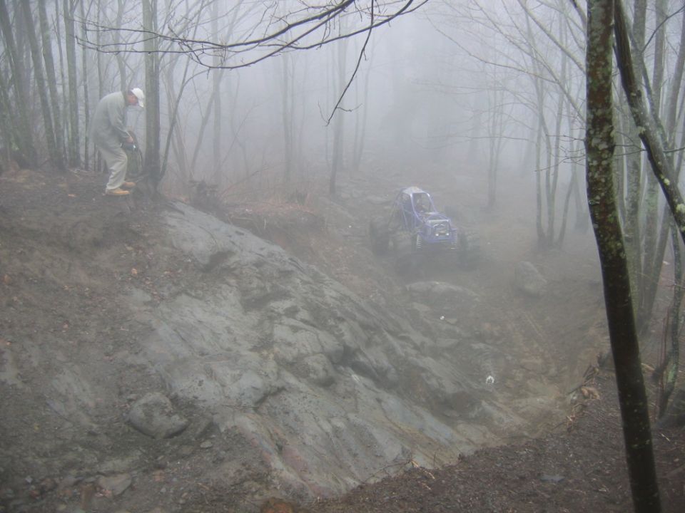 Off-road vehicle navigating a steep, muddy, wooded area with a person standing on the bank above