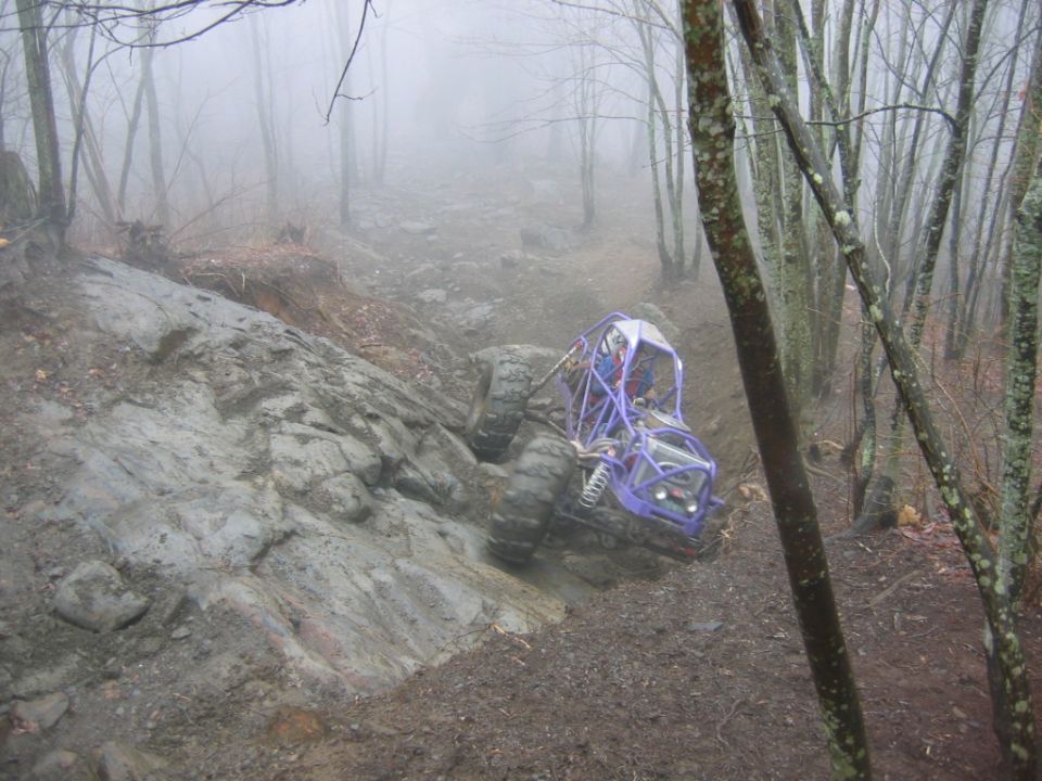 Purple off-road buggy with large tires navigating a steep, rocky, and muddy trail in a foggy forest