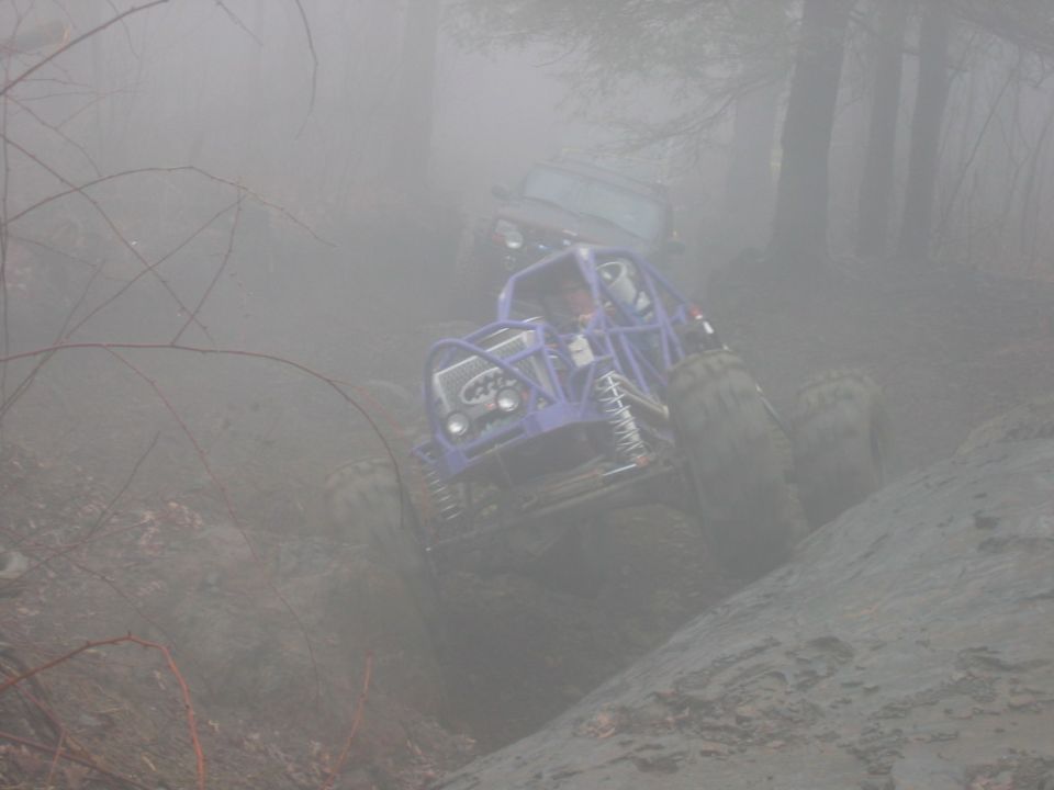 Purple off-road buggy with roll cage climbing a muddy incline, another vehicle visible behind in fog