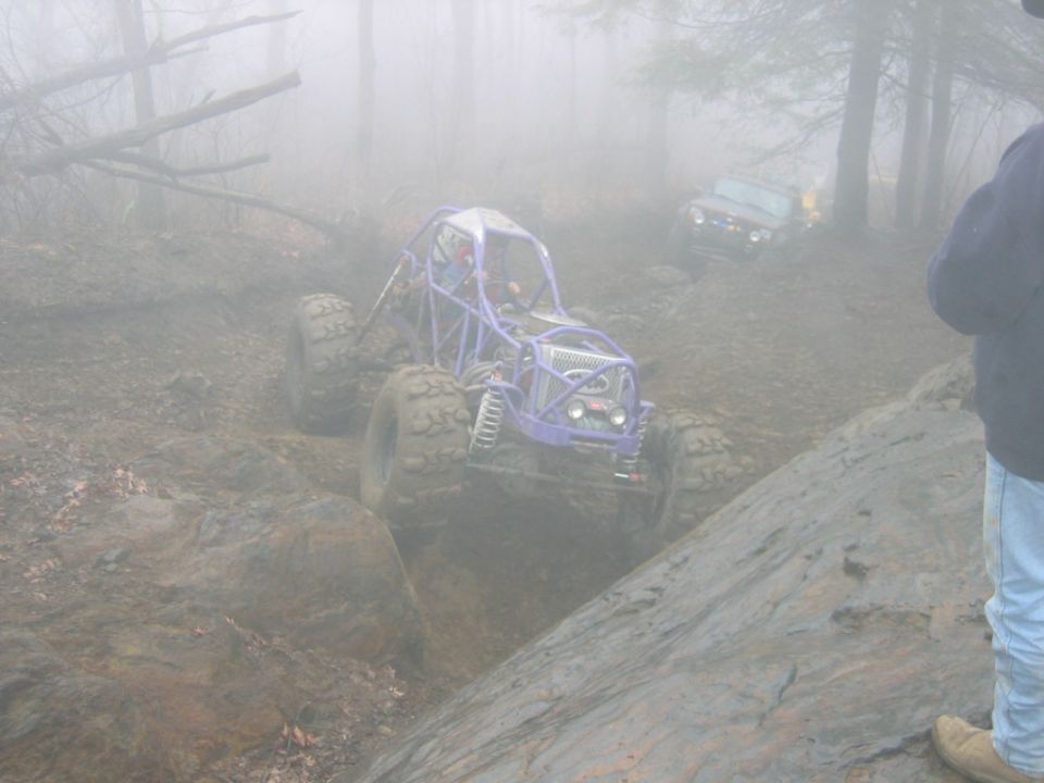 Purple off-road buggy with large tires navigates a muddy, rocky trail in foggy conditions. Another vehicle is visible in the background