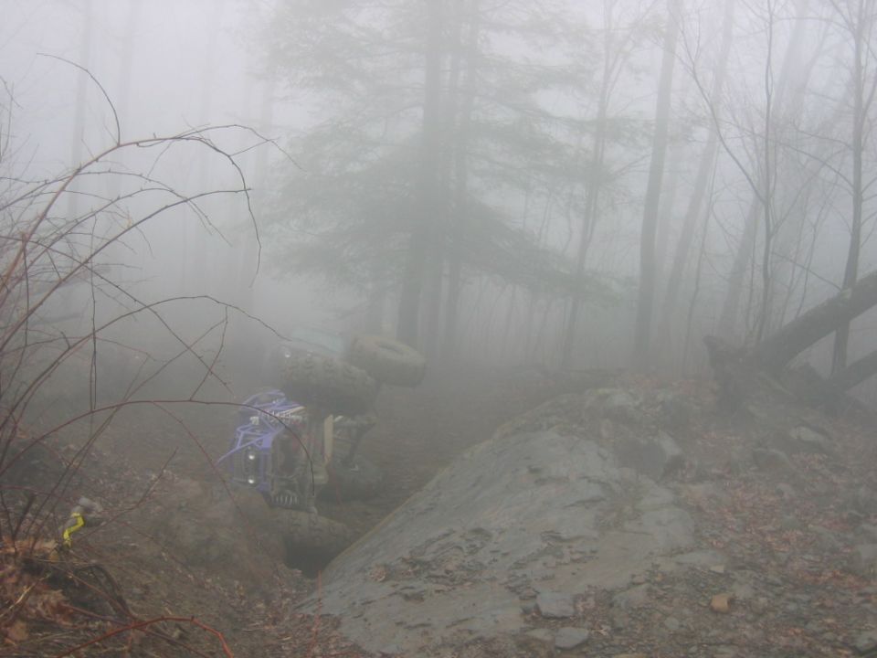 Off-road vehicle tipped on its side in a muddy ditch with trees in the background