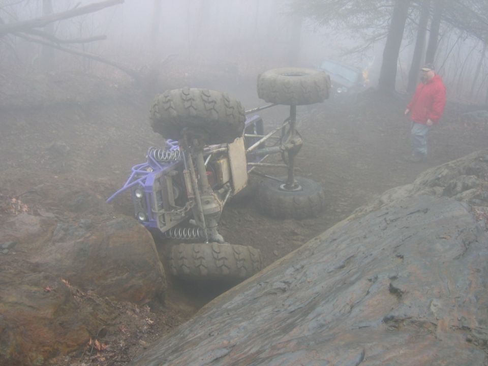 Off-road buggy flipped on its side in a muddy, foggy wooded area. Another vehicle is visible in the background