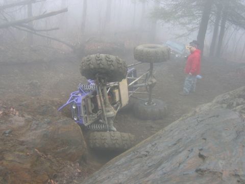 1992 Ford Mustang - An overturned off-road buggy lies on its side in a muddy ditch, with two onlookers observing the scene in a foggy environment.