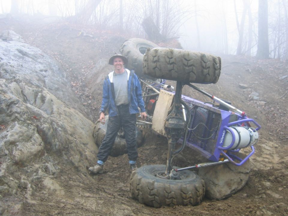 Off-road buggy on its side in a muddy, wooded area with a person standing nearby