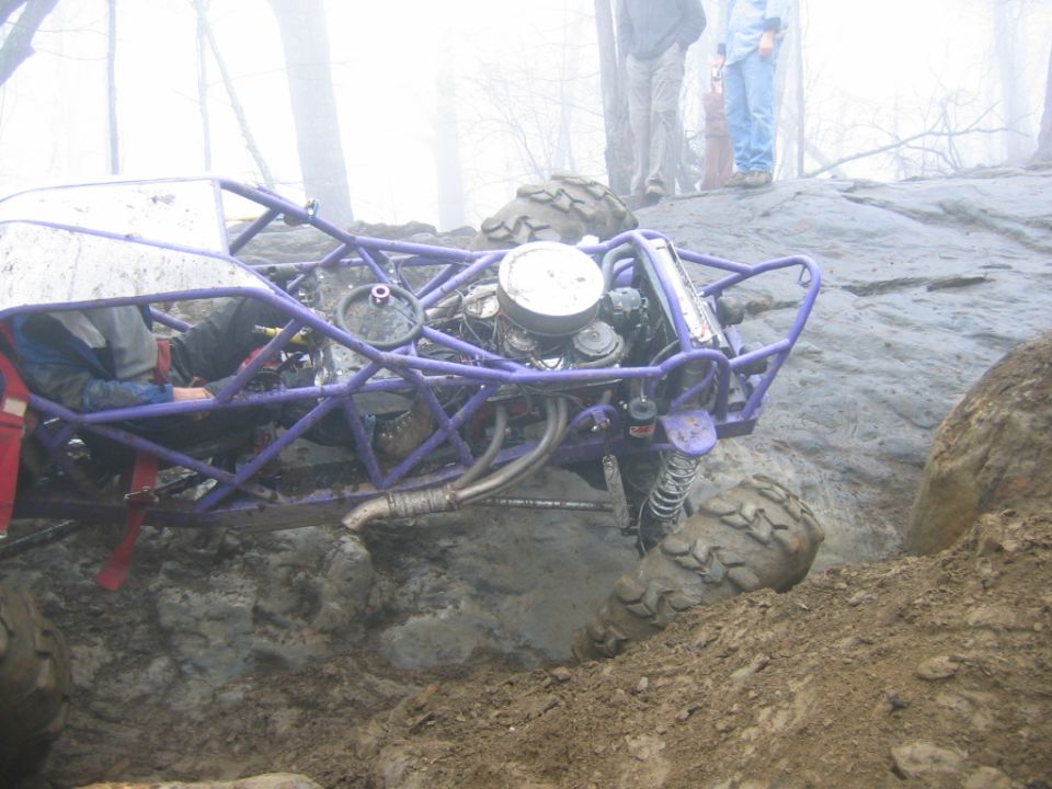 Purple off-road buggy with exposed engine stuck in mud, with people watching in the background