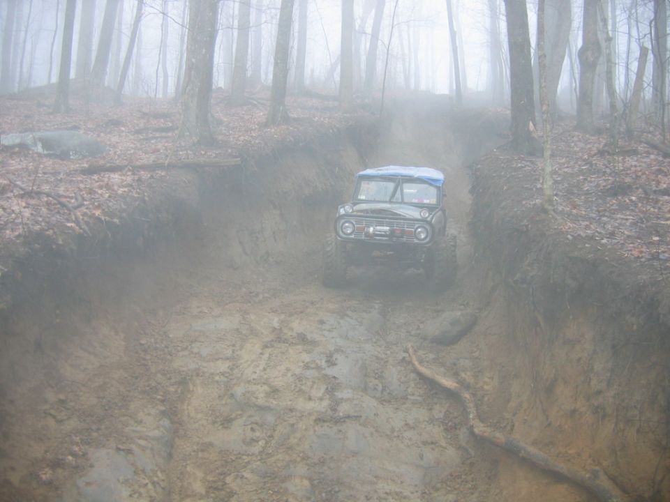 Off-road vehicle with blue tarp on roof driving up a muddy incline in a foggy forest
