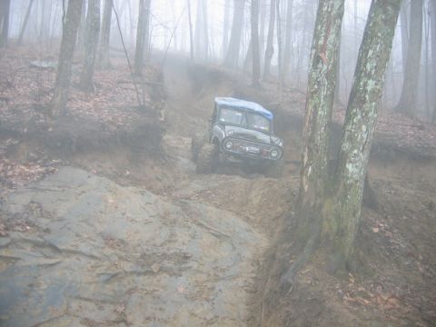 1992 Ford Mustang - An off-road vehicle descends a challenging, muddy incline surrounded by trees in a foggy environment.