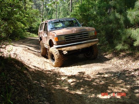 1987 Ford Bronco - A brown 1987 Ford Bronco navigates a muddy, wooded trail with large off-road tires.