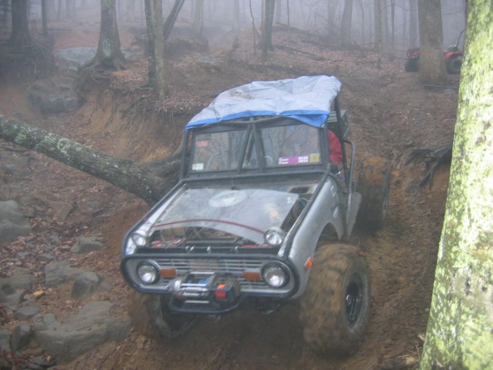 Silver off-road vehicle with a tarp on top, navigating a muddy forest trail with a fallen tree