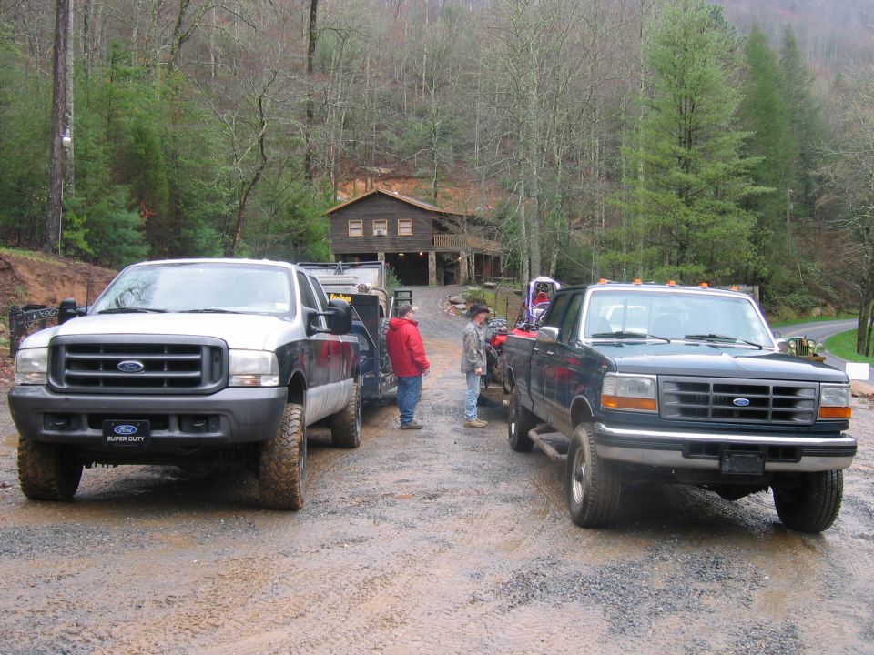 Two Ford F-Series trucks parked on a gravel road, one dark green, one white and black. A house is visible in the background
