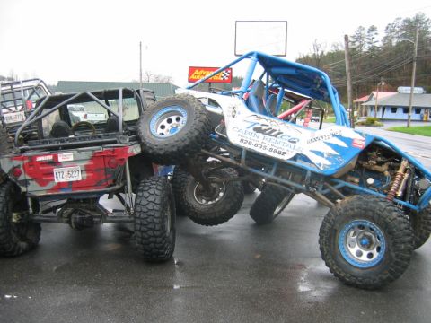 1992 Ford Mustang - Two custom off-road vehicles are positioned closely together on a wet asphalt surface, with one appearing to be on top of the other.