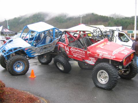 1992 Ford Mustang - Two heavily modified off-road vehicles are parked side-by-side on a wet surface, with a white van and tents in the background.