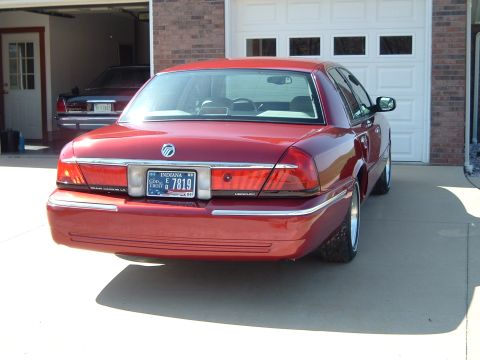 1999 Mercury Grand Marquis - Rear view of a red 1999 Mercury Grand Marquis parked outside a garage with another vehicle inside.
